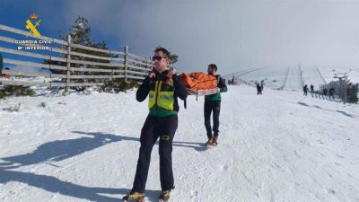 Dos rescatistas en una ladera nevada transportan a hombros una camilla de rescate naranja en un día despejado de montaña.