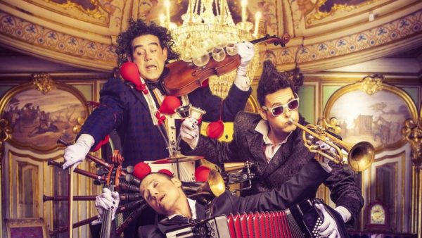 Tres músicos en un entorno palaciego realizan una presentación cómica; uno toca un violín con copas encima, otro una trompeta con gafas de sol y el tercero yace en el suelo con un acordeón.