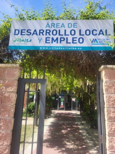Entrada principal al Área de Desarrollo Local y Empleo de Collado Villalba. Un gran cartel informativo corona una puerta metálica rodeada de vegetación.