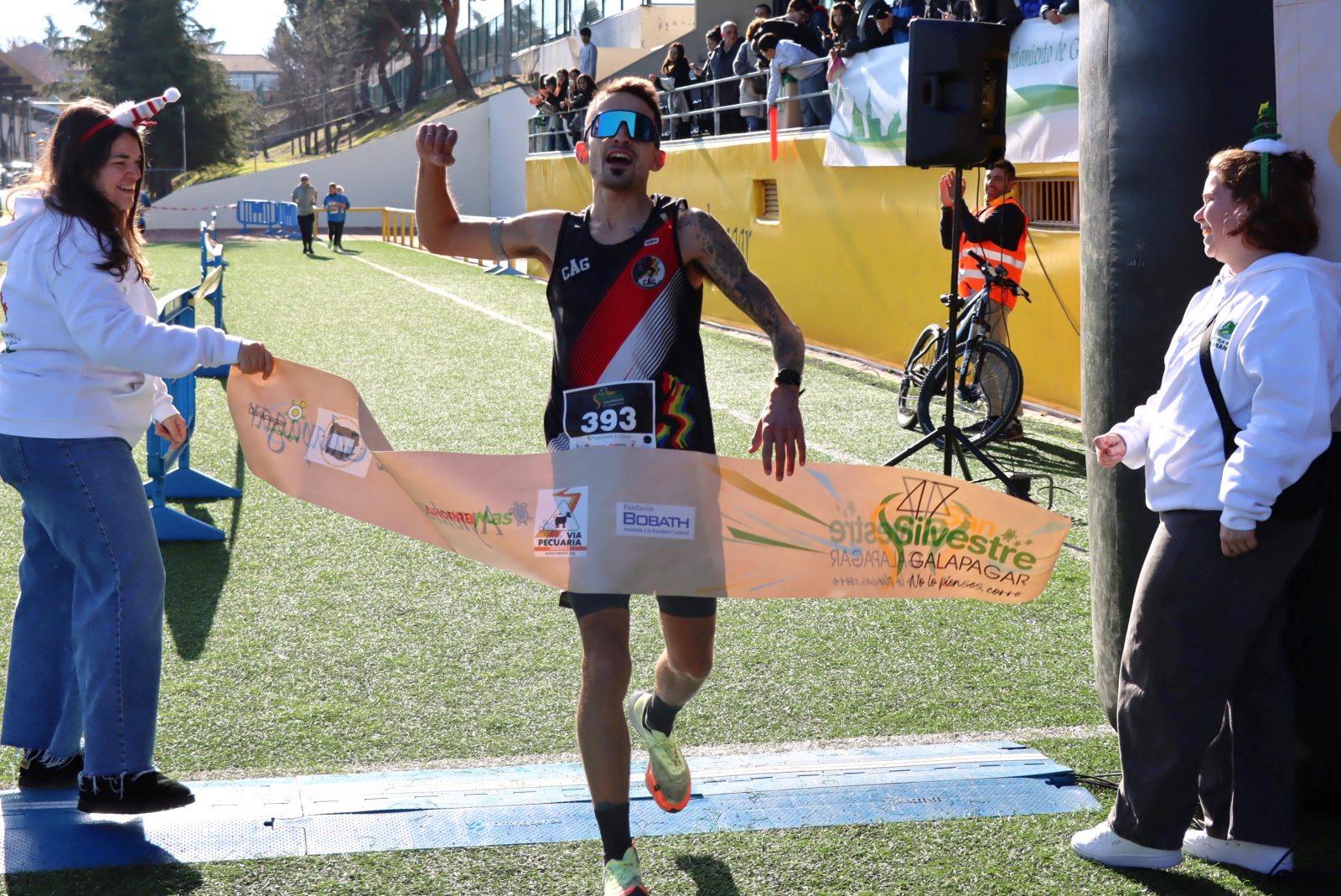 Un corredor con gafas de sol cruza la meta de la San Silvestre de Galapagar rompiendo la cinta con el brazo en alto