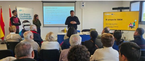 Un grupo de personas mayores y participantes asisten a una presentación del proyecto iDEM en una sala con pantallas informativas y banderas oficiales.