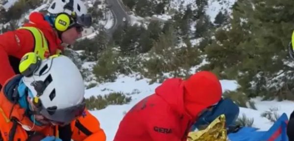 Tres rescatistas con cascos blancos y chaquetas rojas y naranjas trabajando en una ladera nevada sobre una persona herida.