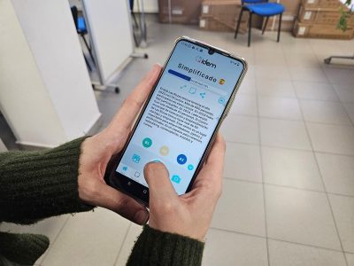 Primer plano de unas manos sosteniendo un teléfono móvil que muestra la aplicación iDEM con un texto simplificado mediante inteligencia artificial y opciones de niveles de lectura.