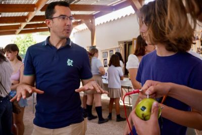 Un adulto con gafas y polo azul oscuro con un logotipo bordado, explica algo gesticulando con las manos a varios jóvenes.