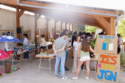 Numerosos adultos y niños conversan y observan proyectos en las mesas dispuestas a lo largo de la zona cubierta.