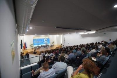 Vista general de un auditorio lleno de gente sentada en butacas azules, presenciando una conferencia