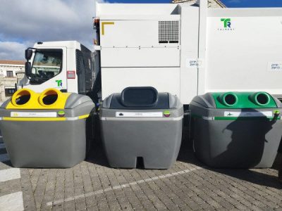 Tres contenedores de reciclaje de tamaño mediano (iglús) alineados en primer plano, con tapas de diferentes colores: amarillo (envases), gris oscuro (resto/basura general) y verde (vidrio).