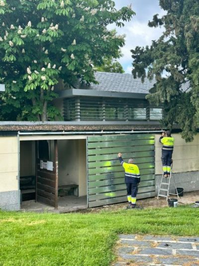 Dos hombres con uniformes de trabajo (pantalones azules y chalecos reflectantes amarillos) pintando una valla metálica con lamas horizontales
