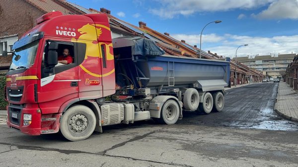 Un gran camión articulado rojo maniobra en una calle residencial, con un tramo de asfalto fresco recién extendido visible detrás del vehículo.