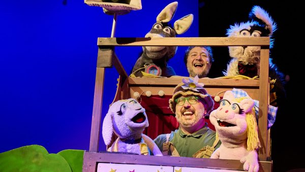 Dos actores sonrientes actúan en un escenario junto a grandes y coloridos títeres de animales como un burro, un perro y una cerdita.