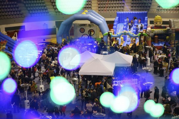 Vista aérea de un polideportivo lleno de familias y grandes atracciones hinchables azules, con luces brillantes desenfocadas en primer plano.