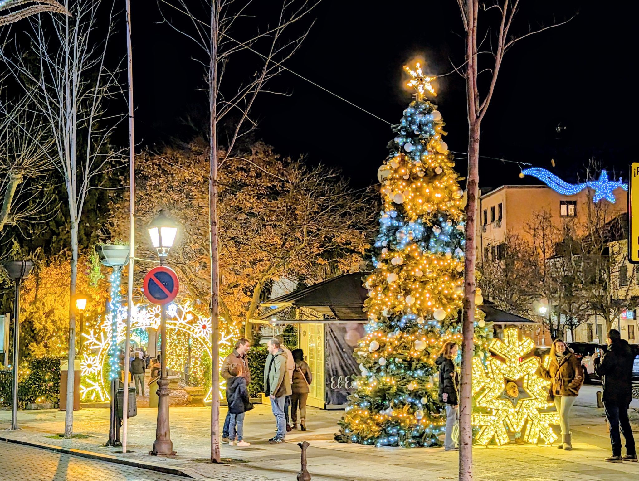 Gran árbol de Navidad exterior decorado con luces azules y doradas junto a un cartel y gente reunida.