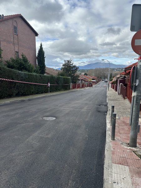 Vista de una calle residencial vacía con asfalto nuevo y oscuro, con chalets a los latos y con montañas al fondo bajo un cielo nublado.