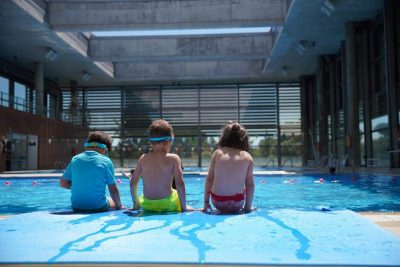 Tres menores sentados de espaldas al borde de una piscina cubierta, preparándose para entrar al agua en un entorno de actividades de verano.