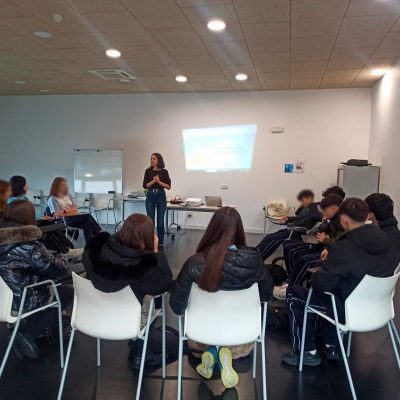 Una orientadora de pie frente a una proyección en una sala con estudiantes de Las Rozas sentados en semicírculo durante los talleres.