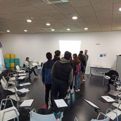 Estudiantes de Las Rozas participan en una dinámica de grupo de pie en el centro de una sala, rodeados de sillas con pupitres.