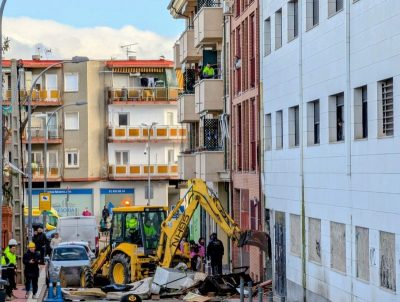 Una excavadora amarilla retira escombros y muebles acumulados en la entrada de un edificio de ladrillo y fachada blanca.