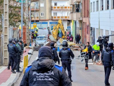 Vista de una calle estrecha en Collado Villalba durante el desalojo. En primer plano, varios agentes de la Policía Local de espaldas acordonan la zona.