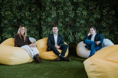 Tres personas, dos hombres y una mujer, sentados en pufs amarillos y blancos en una zona de descanso con césped artificial y una pared vegetal de fondo.