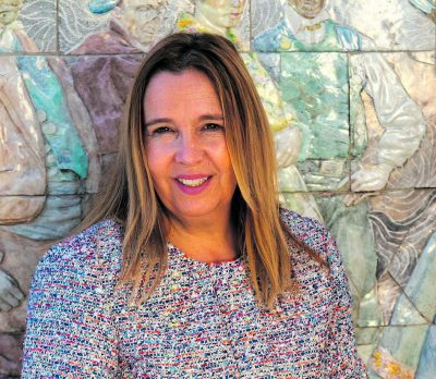Primer plano de Victoria Barderas, alcaldesa de Hoyo de Manzanares, sonriendo frente a un mural de azulejos decorativos.