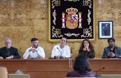 Mesa de sesión en el Ayuntamiento con varios concejales y representantes políticos sentados ante el escudo de España.