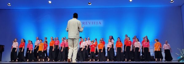 Imagen de un numeroso coro infantil y juvenil sobre el escenario, vestidos con camisetas de colores naranja, rosa y rojo con pantalones negros.