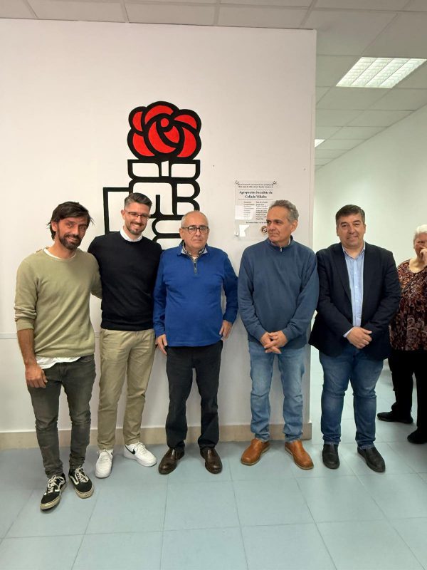 Grupo de asistentes al acto del 90º aniversario del PSOE de Collado Villalba posando frente al logotipo del partido en la sede local.
