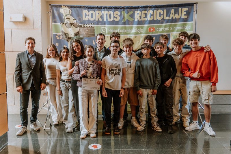 Un grupo de jóvenes y adultos posan sonrientes delante de una lona del proyecto "Cortos x Reciclaje". Una joven en el centro sostiene una claqueta de cine.
