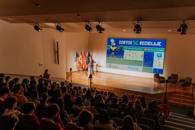 En el escenario, un ponente habla junto a una gran pantalla que muestra el logo del proyecto "Cortos x Reciclaje".