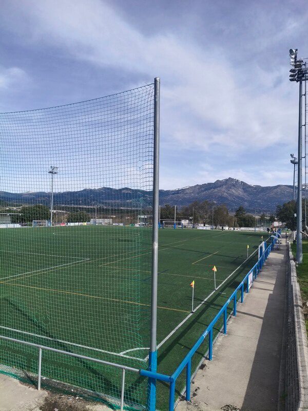 Vista general del campo de césped artificial con redes de protección y la montaña al fondo bajo un cielo despejado.