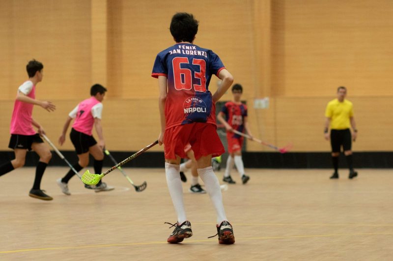 Varios jóvenes juegan un partido de floorball en un pabellón deportivo con sticks y protecciones ante un árbitro.