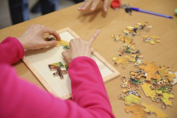 Vista cenital de las manos de una persona con jersey rosa que encaja piezas de un puzle de madera sobre una mesa.