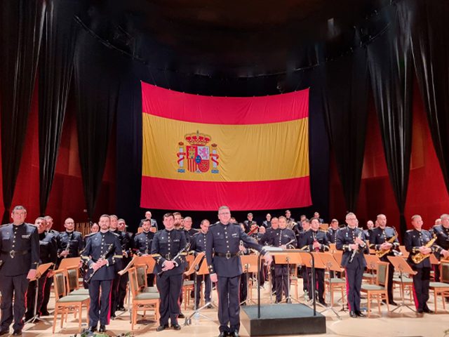 La Unidad de Música de la Agrupación de Infantería de Marina de Madrid posa en un escenario bajo una gran bandera de España.