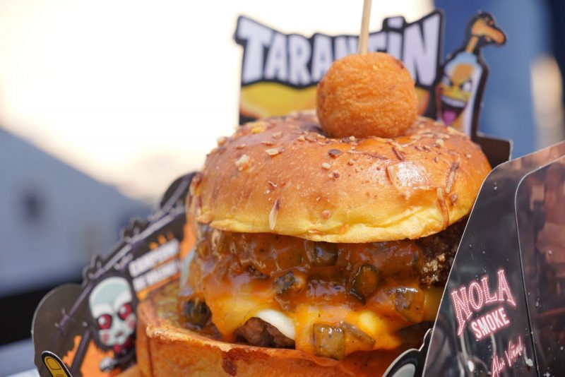 Primer plano de una hamburguesa gourmet con salsa naranja, queso fundido y una croqueta pinchada encima con un palillo.