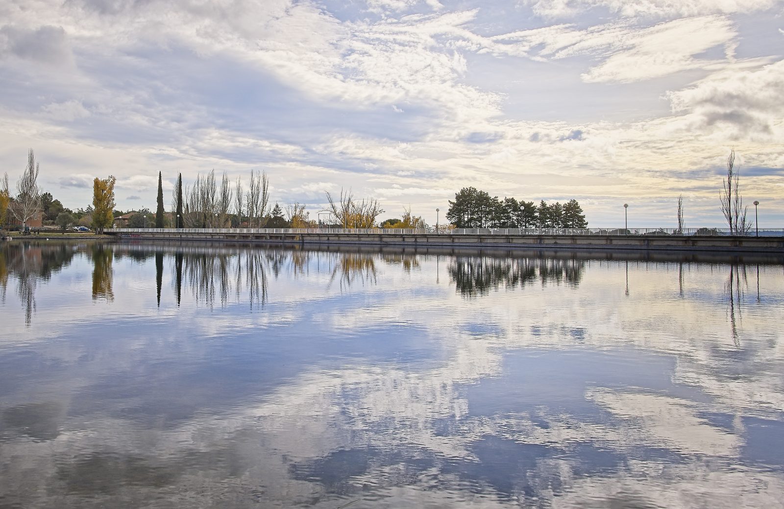 Paisaje del embalse de Los Rosales en Galapagar con árboles y nubes reflejados en el agua tranquila.