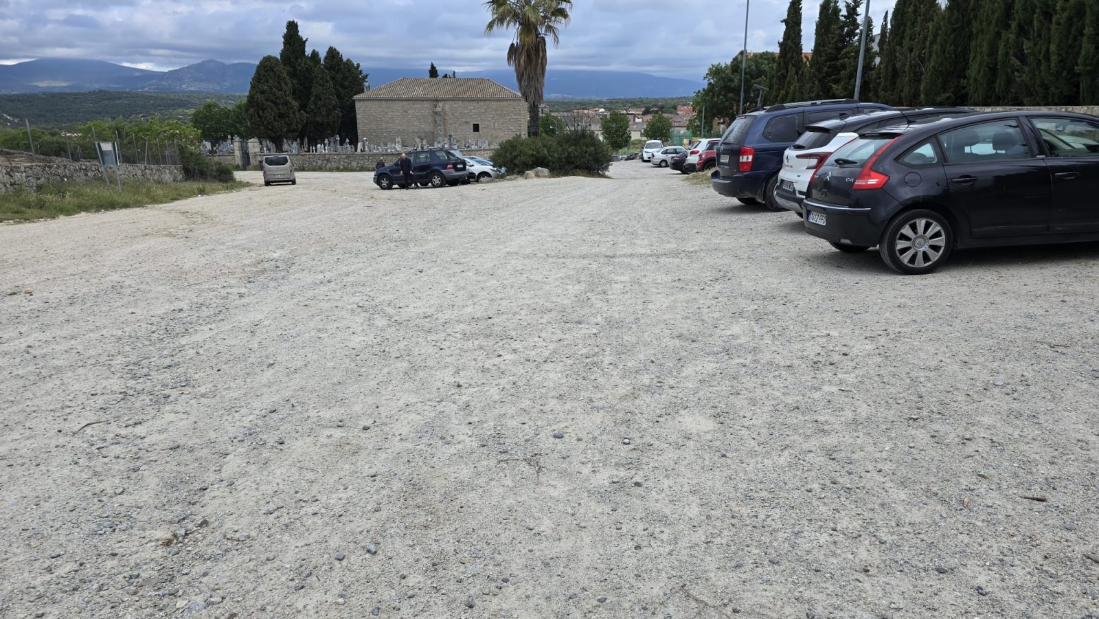 Vista de un aparcamiento de tierra con varios coches estacionados y un edificio de piedra al fondo.