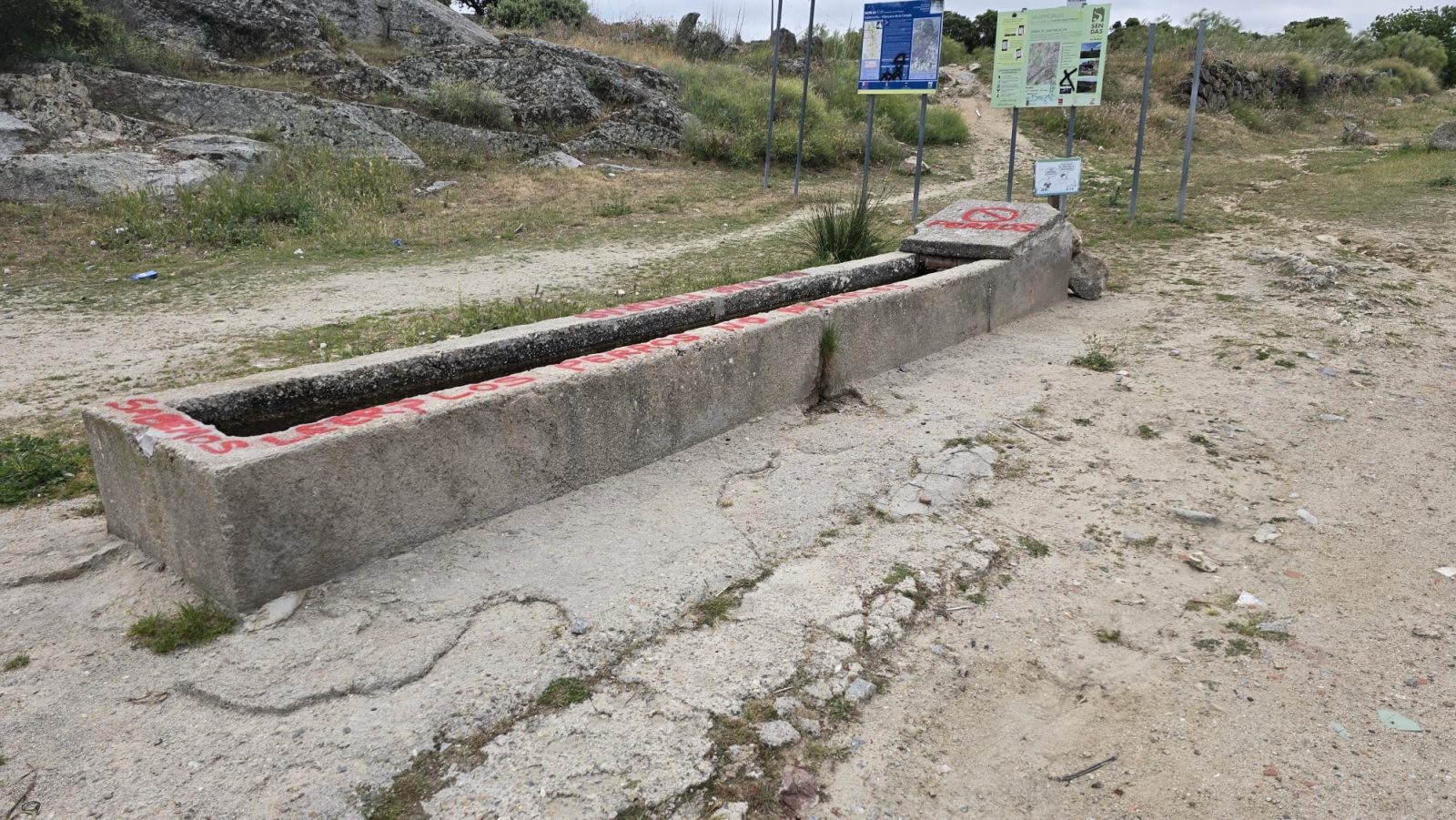 Un abrevadero de piedra alargado en un entorno natural con carteles informativos y rocas al fondo.