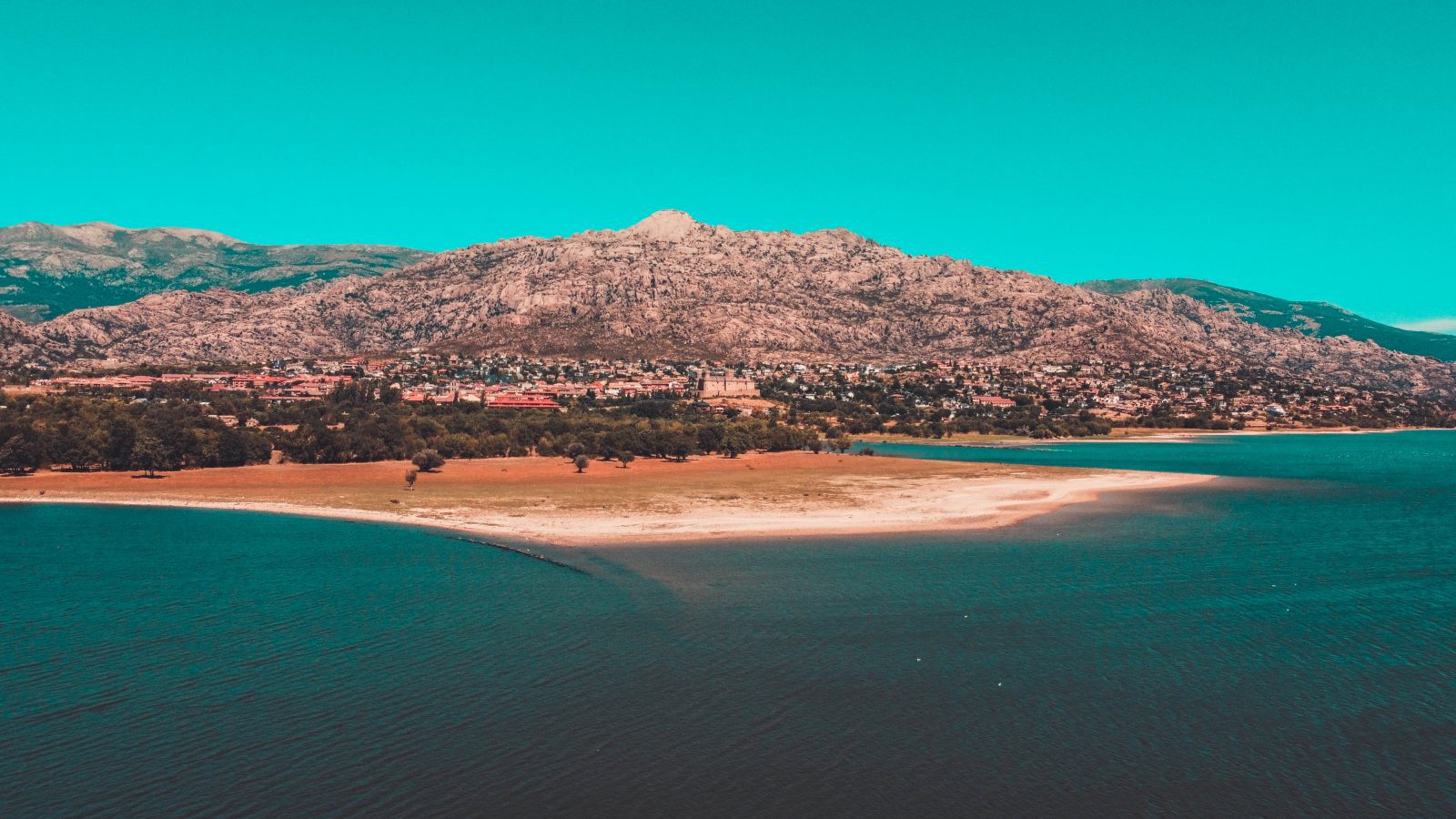 Vista panorámica del embalse de Santillana en primer plano, con el pueblo de Manzanares el Real y la Pedriza al fondo bajo un cielo azul.