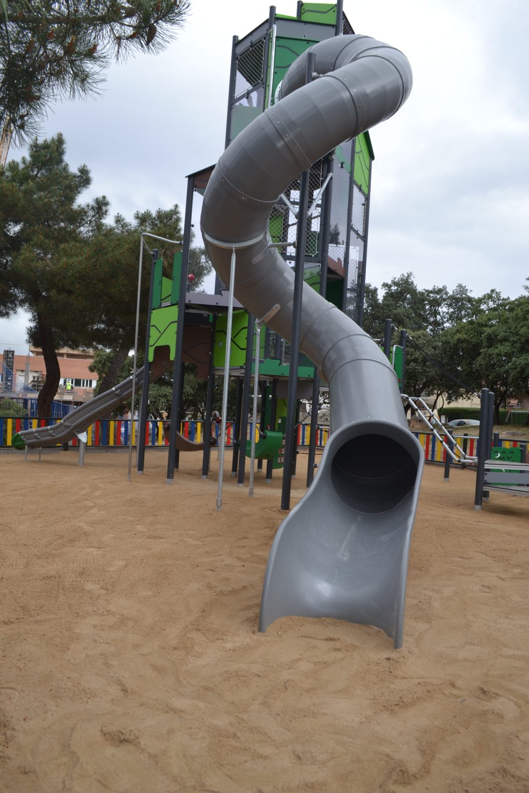 Gran tobogán gris en forma de espiral conectado a una torre de juegos de 9 metros de altura sobre suelo de arena.