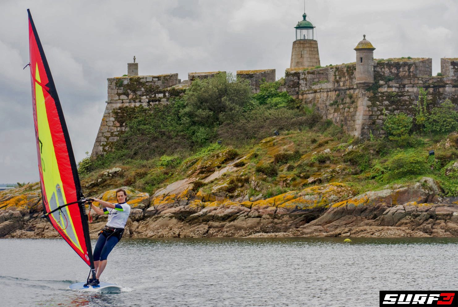 Elena Boyé practica windsurf en la ría frente a una fortificación antigua de piedra con un faro.