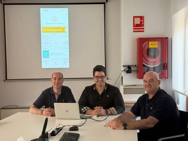 Tres hombres sentados a una mesa con un portátil frente a una pantalla que proyecta la interfaz de una aplicación móvil.