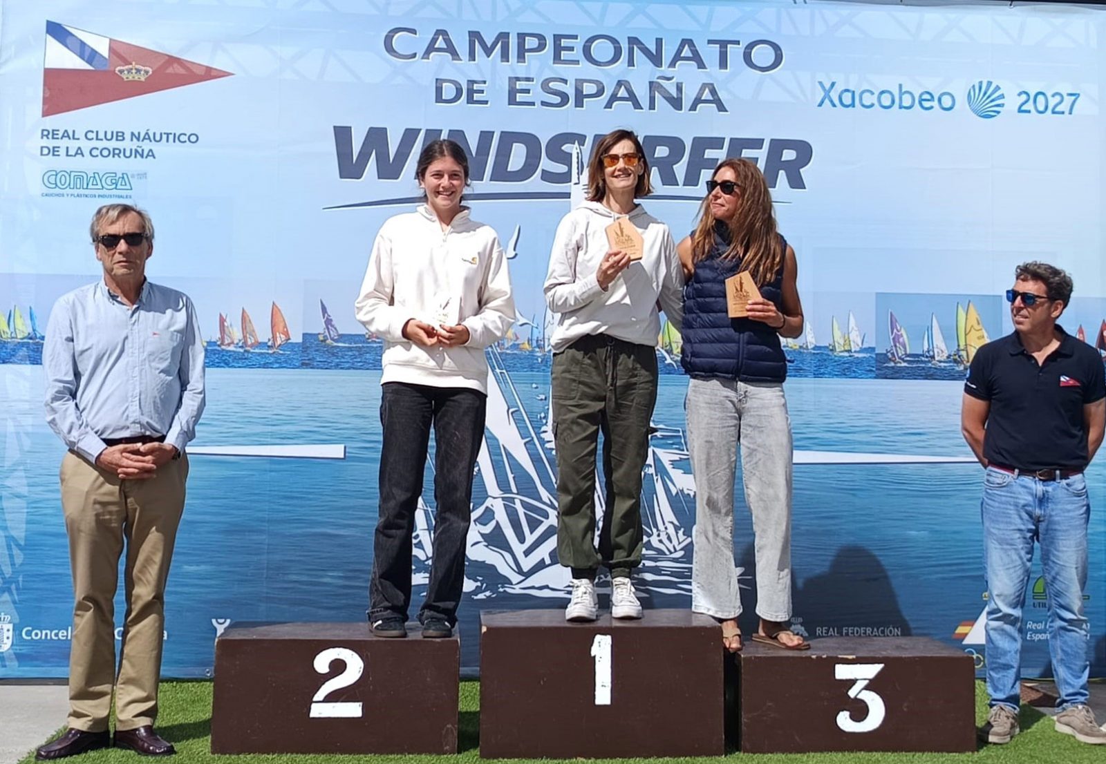 Tres deportistas en un podio con trofeos frente a un cartel del Campeonato de España de Windsurfer.