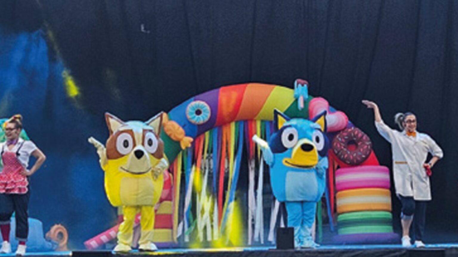 Dos actores con disfraces de personajes infantiles y dos animadoras actúan en un escenario frente a un arco de colores.