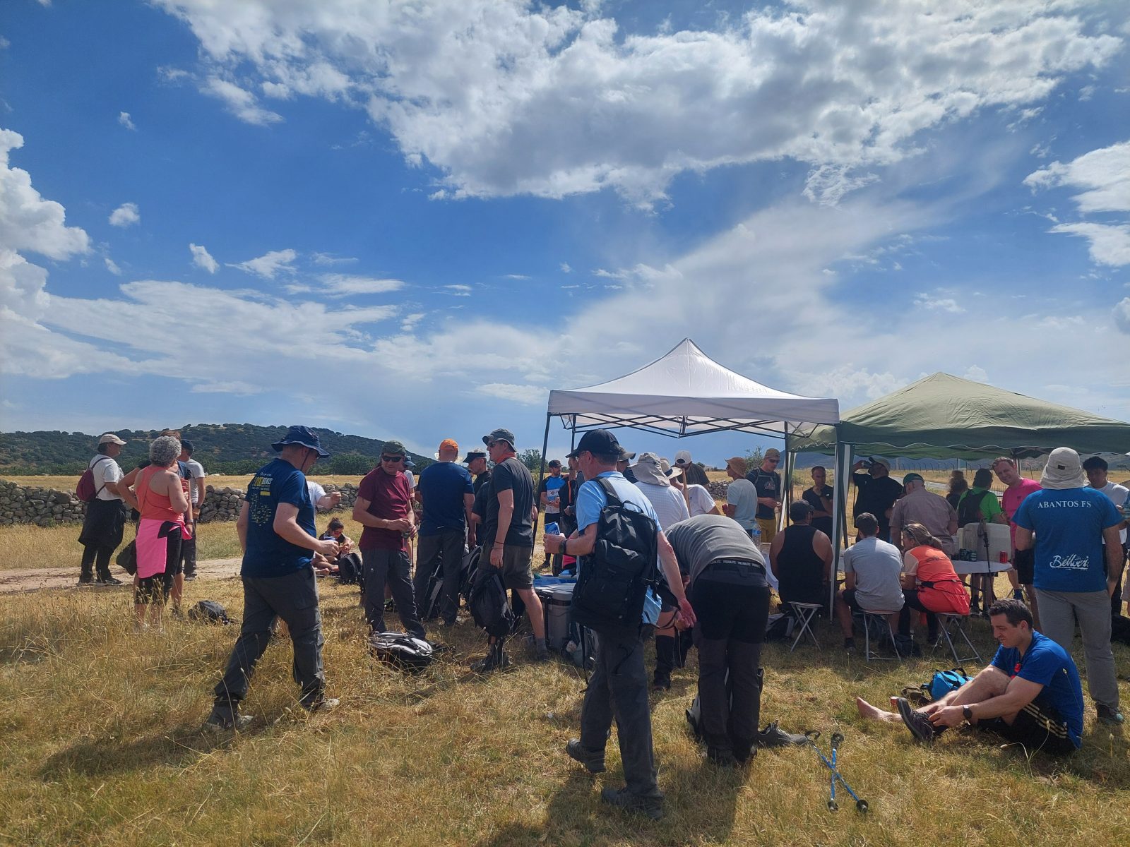 Senderistas descansando y congregados alrededor de unas carpas blancas instaladas en una zona de pasto seco bajo un cielo con nubes.