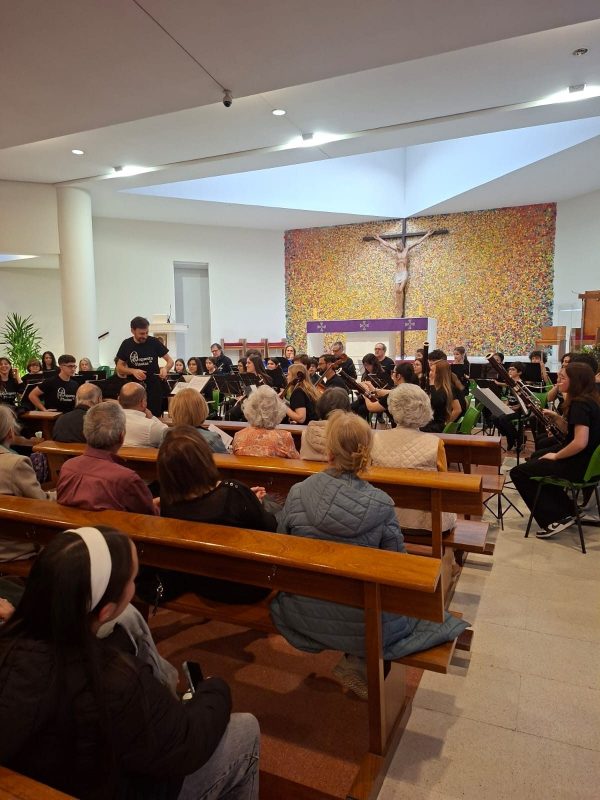 Una orquesta juvenil con instrumentos de viento actúa frente al público sentado en los bancos de una iglesia moderna.