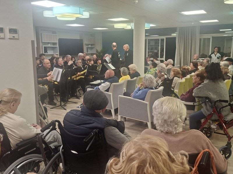 Un grupo de músicos toca instrumentos de viento para un público de personas mayores sentadas en sillas y sillas de ruedas.