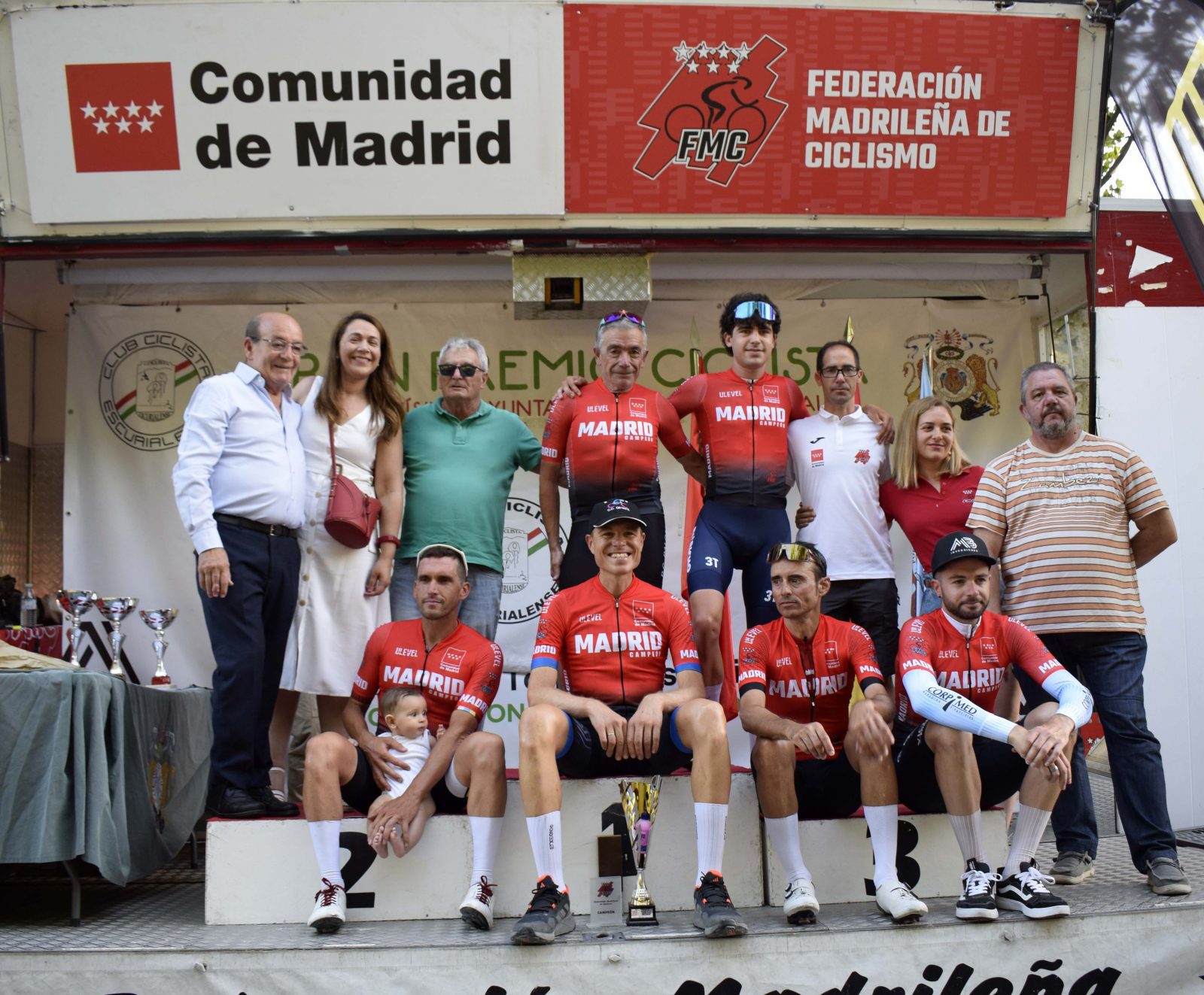 Ciclistas con maillots rojos de campeones de Madrid posan en el podio junto a autoridades y organizadores del evento.