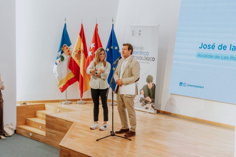 El alcalde de Las Rozas, José de la Uz, habla ante un micrófono en un escenario junto a una mujer y cuatro banderas oficiales de fondo.