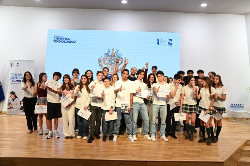 Grupo numeroso de jóvenes posa sonriendo y levantando el pulgar en un escenario con diplomas frente a una gran pantalla azul.