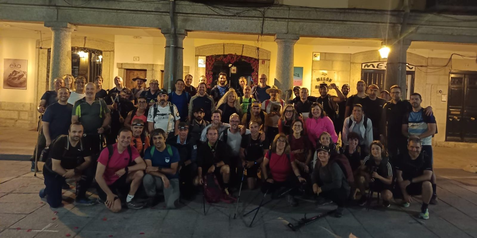 Gran grupo de senderistas posando de noche antes de iniciar la marcha frente a los soportales de una plaza iluminada.